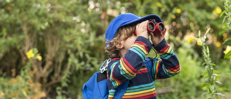 A young person wearing a blue cap and rucksack looking through binoculars